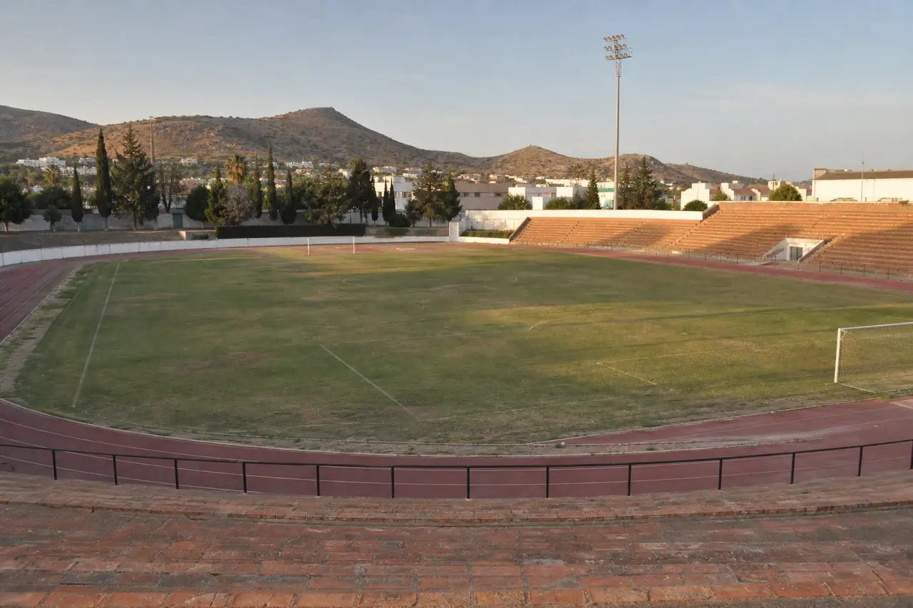 Late-afternoon municipal football stadium in Spain with worn grass, a clay running track and empty terracotta seating
