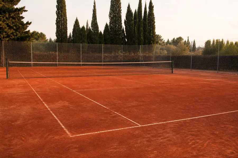 Empty red clay tennis court with cypress trees beyond the fence